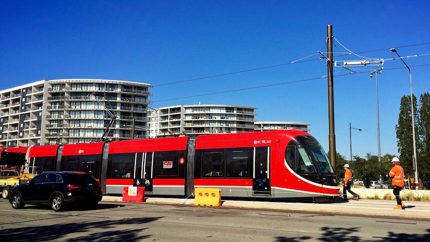 A bright red light rail tram sits idle on the tracks in Canberra with workers nearby.