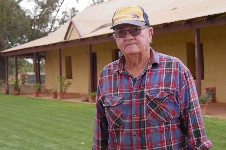 A man in a checked shirt and West Coast Eagles hat stands outside a yellow house. There are pot plants along the verandah.