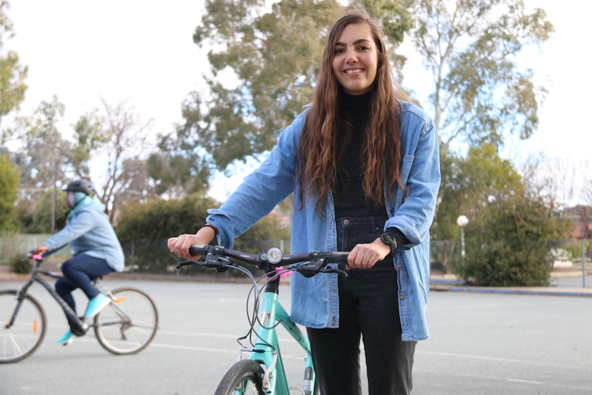 A woman with long brown hair stands with a bike
