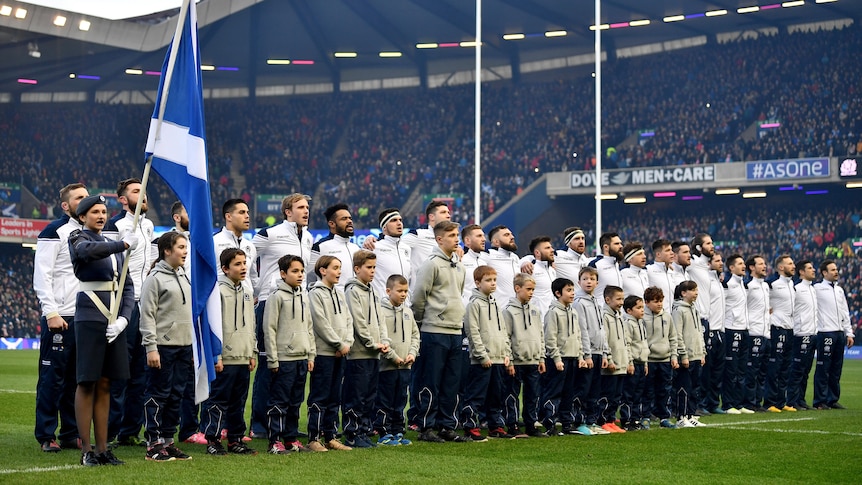 The Scotland team lines up and sings the national anthem in a stadium with a Scottish saltire flag being held near them