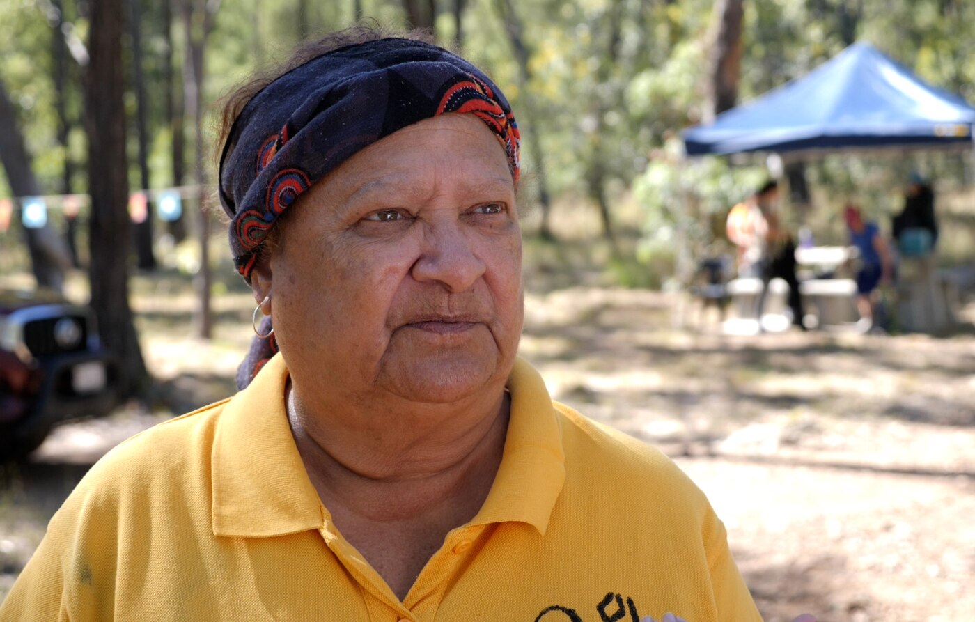 Aunty Sally wearing a headband, yellow shirt, straight face, trees and tent behind.