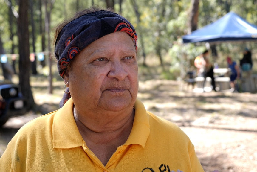 An indigenous woman wearing a headband, yellow shirt, straight face, trees and tent behind.