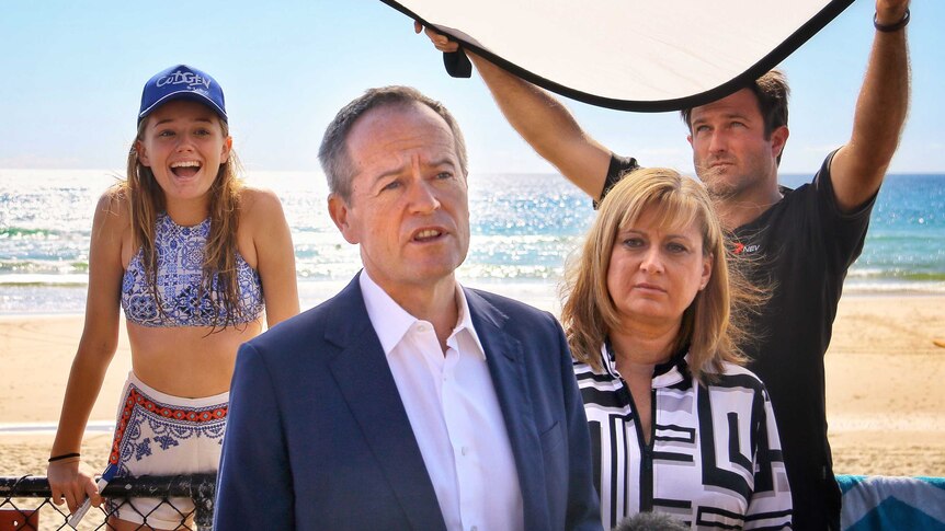 Bill Shorten holds a media conference on the beach as a girl lifts herself up on a fence behind him and smiles broadly.
