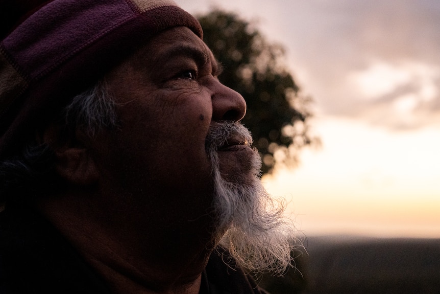 An older Aboriginal man with a weathered face and white beard stands with a concerned expression as the sun rises behind him.