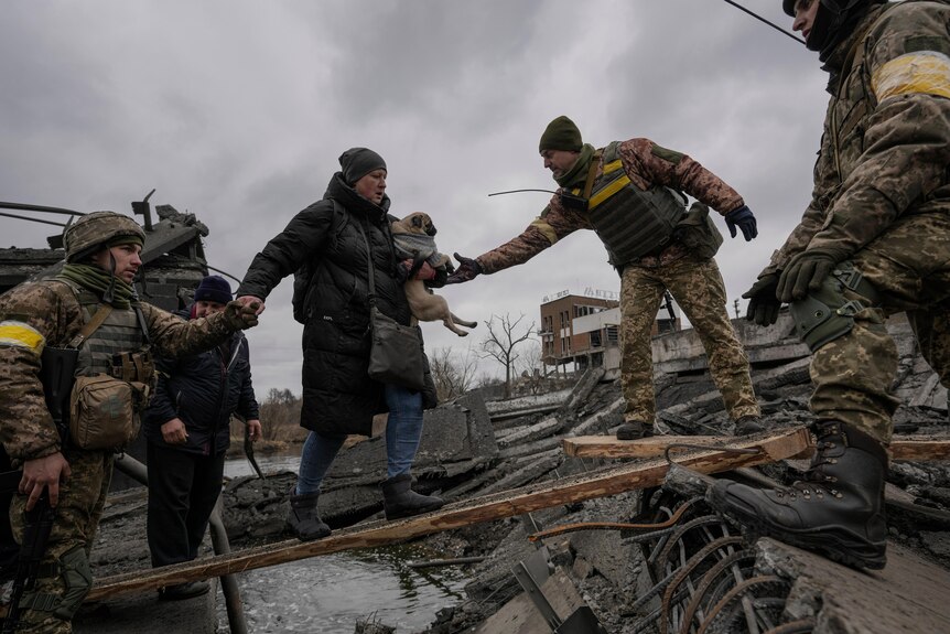 Ukrainian servicemen help a woman carrying small dog cross a river on a wooden plank 