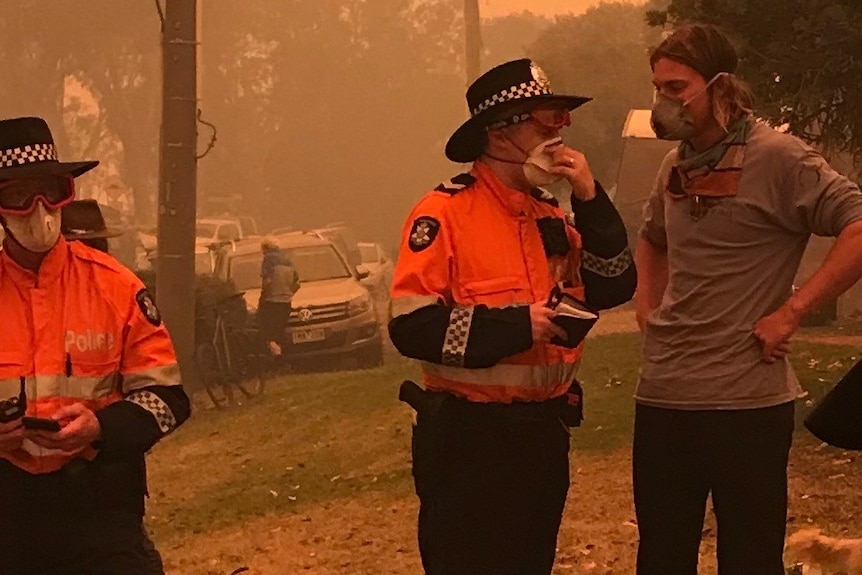 Two police officers and one civilian wearing mask over their nose and mouth talk on a smoky street.