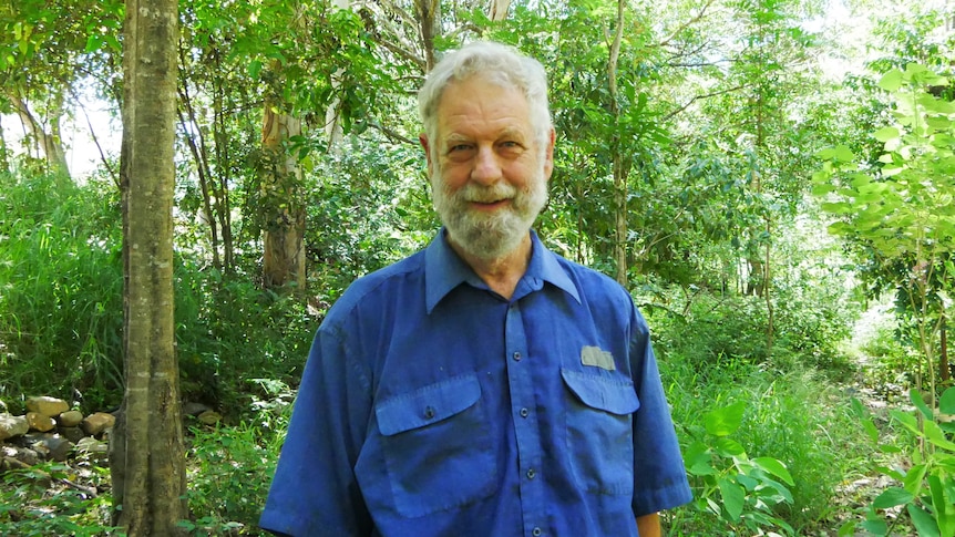 A man with grey hair and a beard wearing a navy work shirt smiles, in the background is bushland