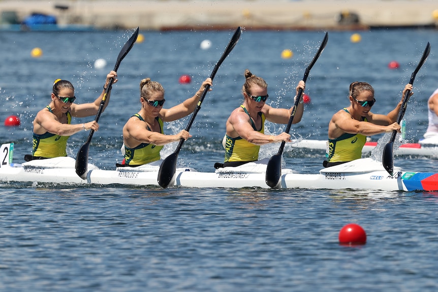 Four women wearing yellow shirts sit in a boat holding oars