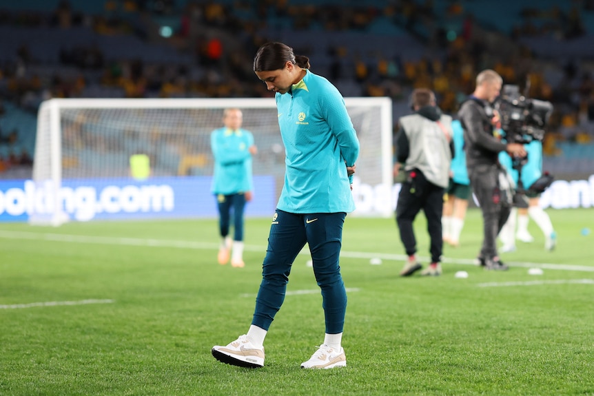A woman wearing a tracksuit of light blue and green walks on a soccer field with a cameraman behind her