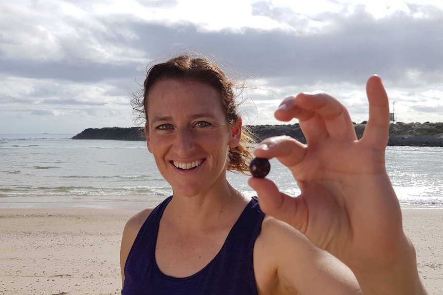 A woman stands on a beach holding a small black marble up to the camera.