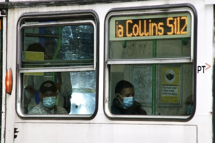 Two people with masks on sit in a tram, photographed through windows from the outside.