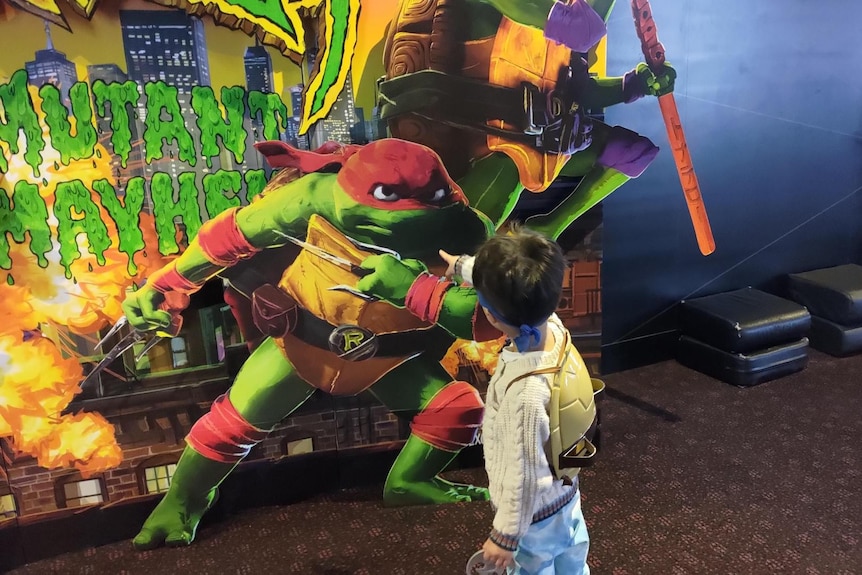 Young boys points at a movie poster with a large ninja turtle on it.