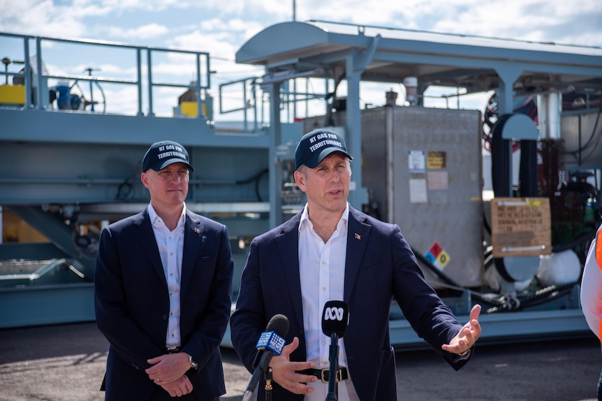 Two men wearing dress shirts, jackets and caps stand in front of a drilling rig