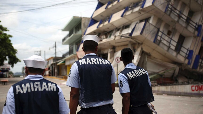 Three men dressed in naval uniforms walk towards a collapsed hotel.