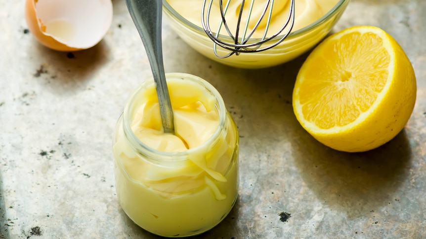A jar of homemade mayonnaise alongside a bowl with a whisk and a lemon.