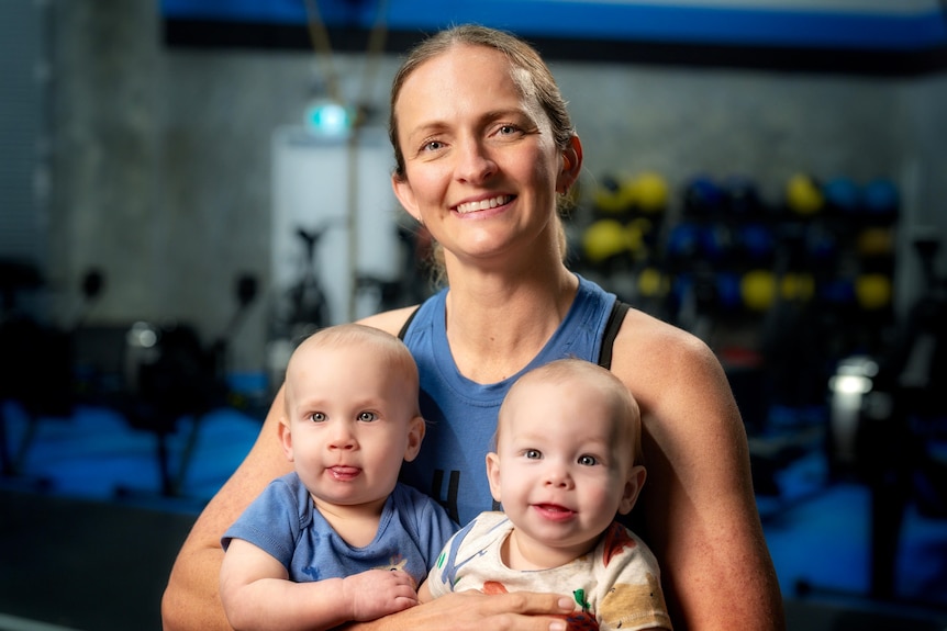 A smiling white woman holding two babies at the gym