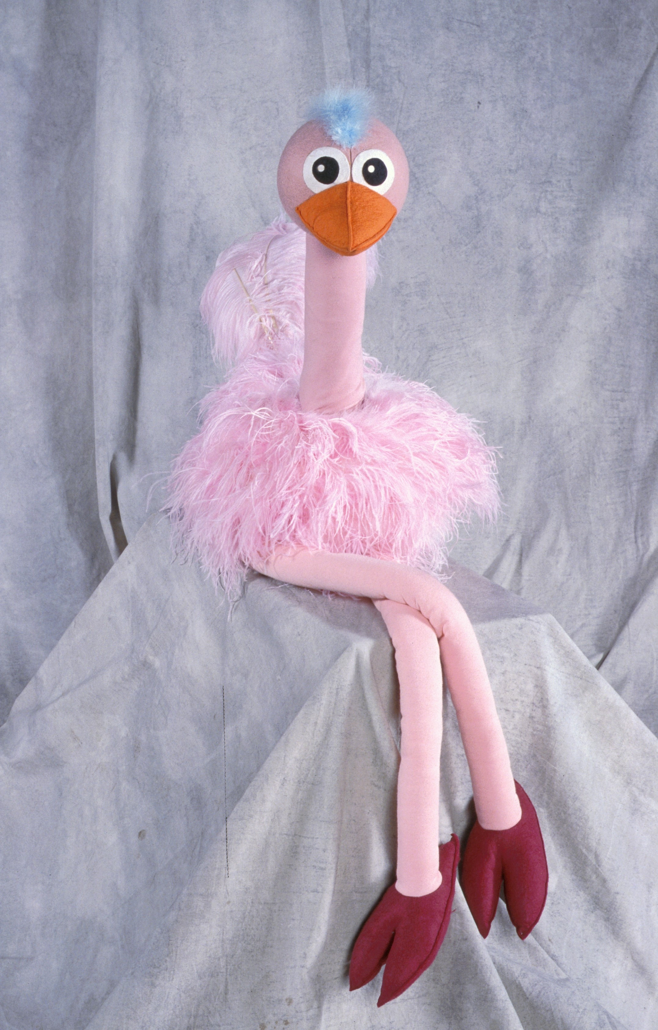 A pink ostrich puppet with a blue mohawk and orange beak is propped in a studio with its legs crossed.
