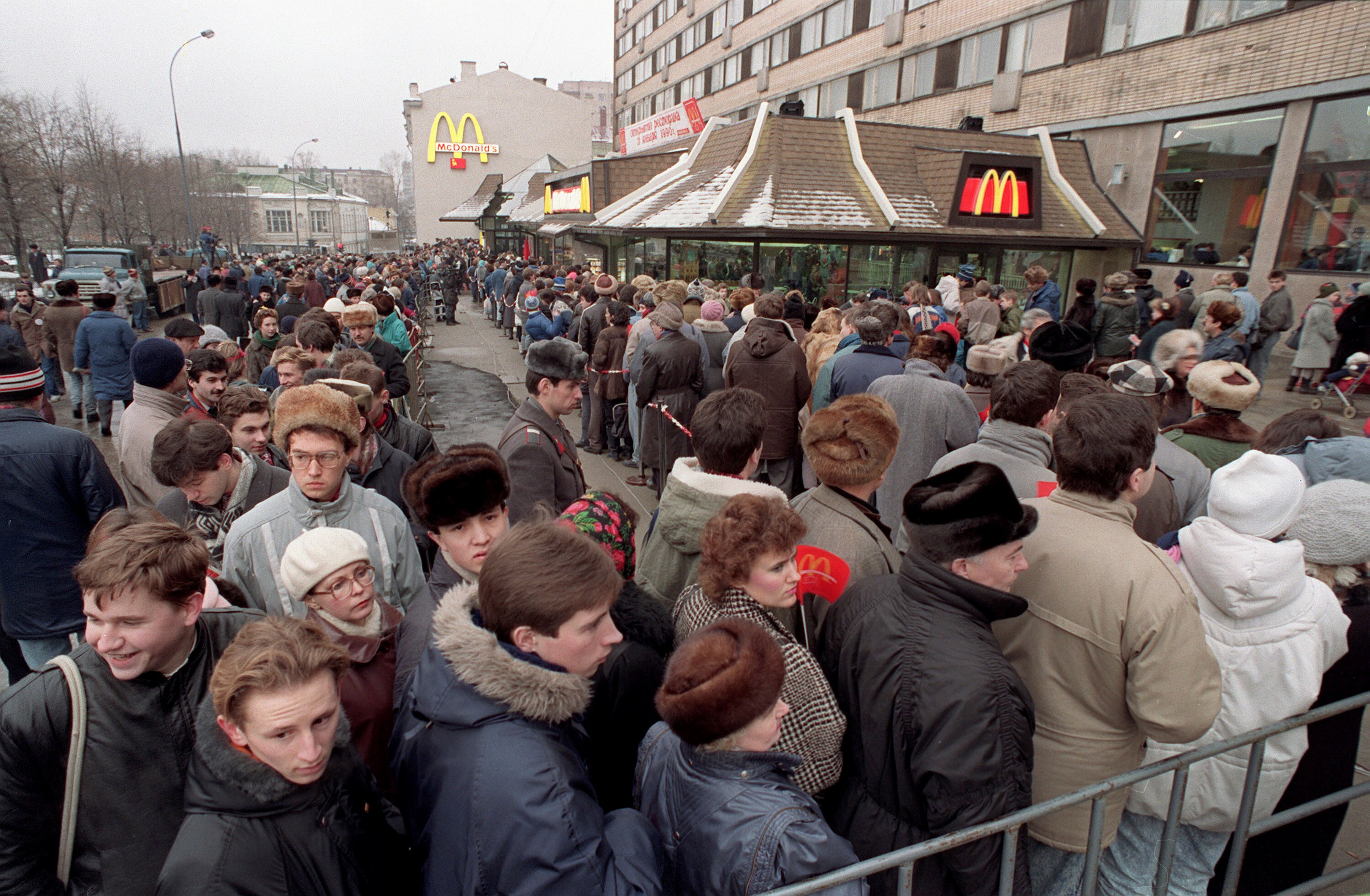 A long queue of people dressed in Soviet-style clothing twists around in front of a building with prominent McDonald's logos.