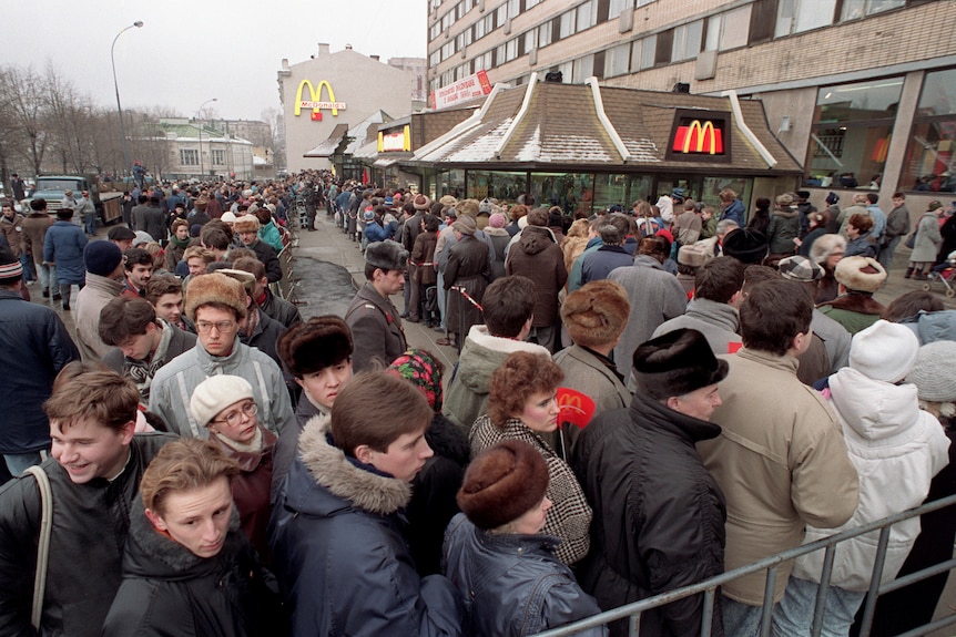 A long queue of people dressed in Soviet-style clothing twists around in front of a building with prominent McDonald's logos.