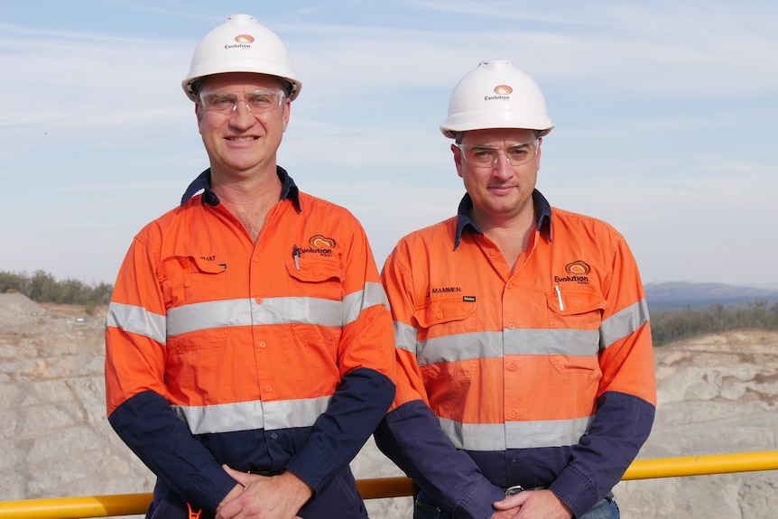 Two men in hi-vis and white hard hats stand in front of a gold mine with blue sky behind them
