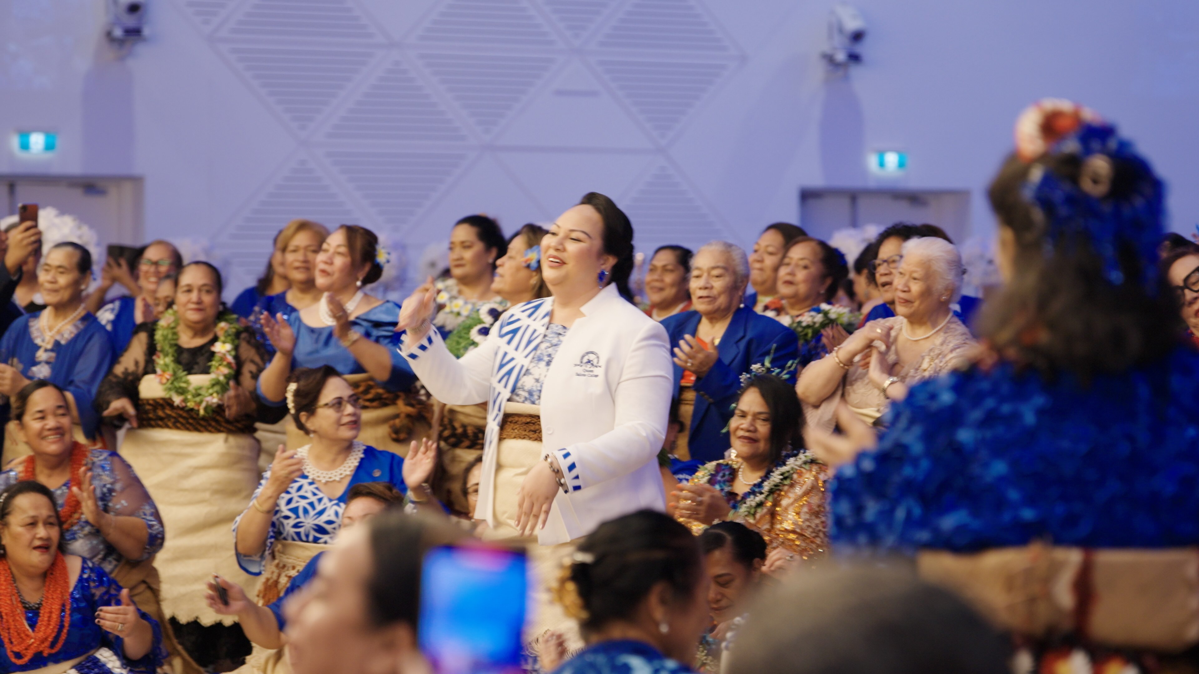 Lady in white jacket with blue patterns and mats around her waist, smiles with her arms up to dance with group of women behind 