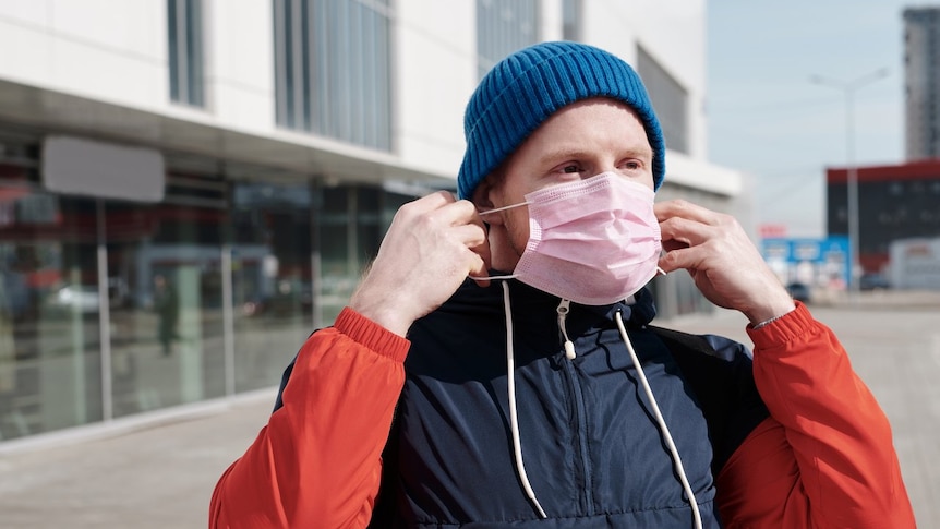 A man sits on a scooter and puts a pink surgical mask on his face. In the background is a white building.