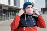A man sits on a scooter putting a pink surgical mask on his face