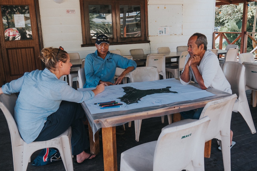 A fair-haired woman in a light blue shirt sits at a table with two Christmas Island men, one wearing a navy blue cap