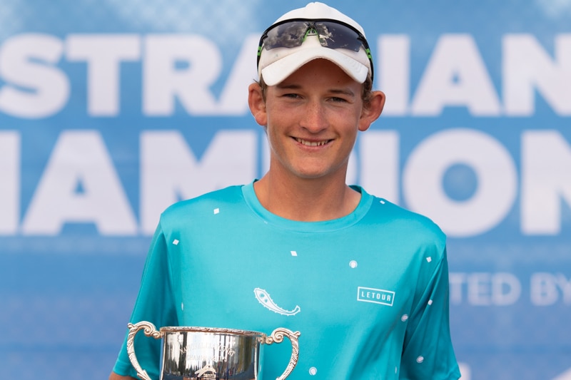 A boy wearing a blue shirt, white cap and black glasses smiles with a trophy.