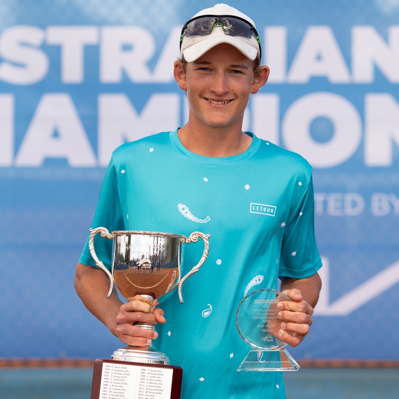 A boy wearing a blue shirt, white cap and black glasses smiles with a trophy.