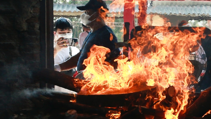 A man wearing a mask and hat walks by a fire blazing in a room.