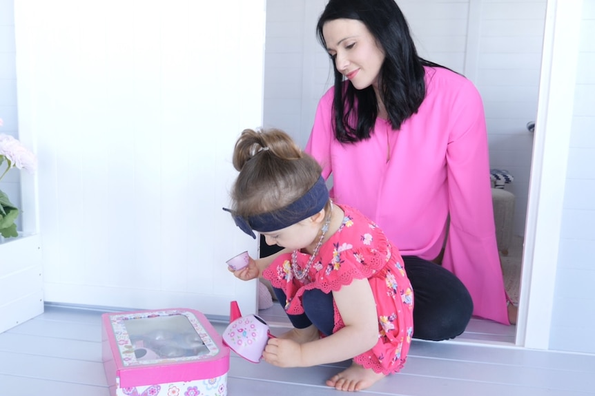 Catherine sits in the doorway of a cubby house and watches Charlotte play with a tea set.