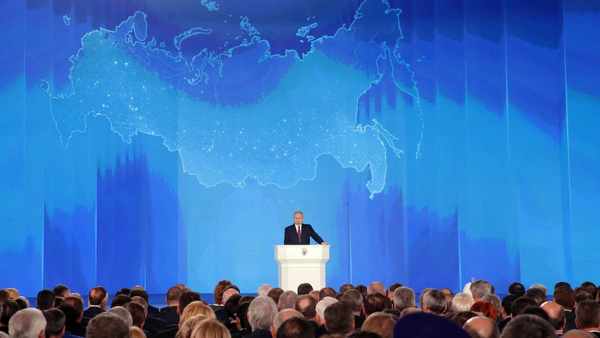 Vladimir Putin address an assembly against the backdrop of a digital map of Russia.