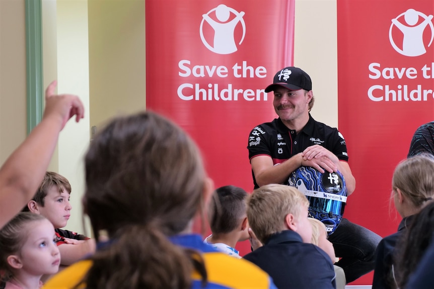 A smiling man holds a racing helmet on his knee while he talks to a group of young kids.
