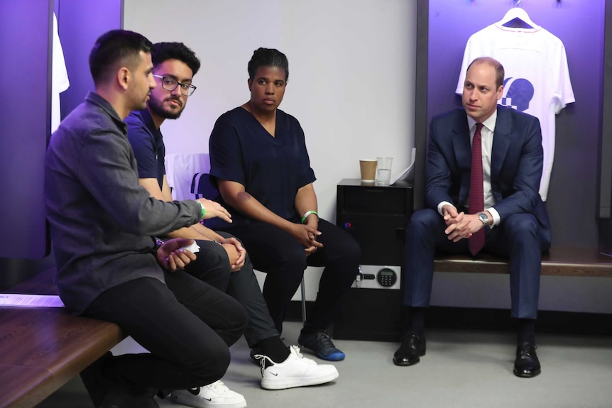 In a purple-hued soccer locker room, Prince William sits on a bench with two men and one woman in conversation.