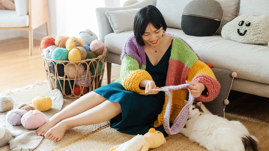 Karen sits while knitting wearing a multicoloured, chunky knit cardigan next to a basket with wool and a fluffy white cat.