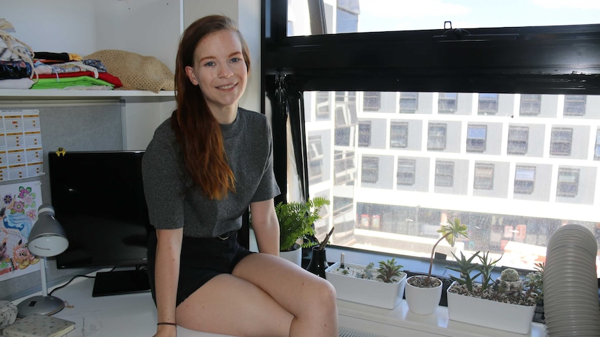 Kaitlyn sits on her desk next to her window, plants alongside her in small pots.