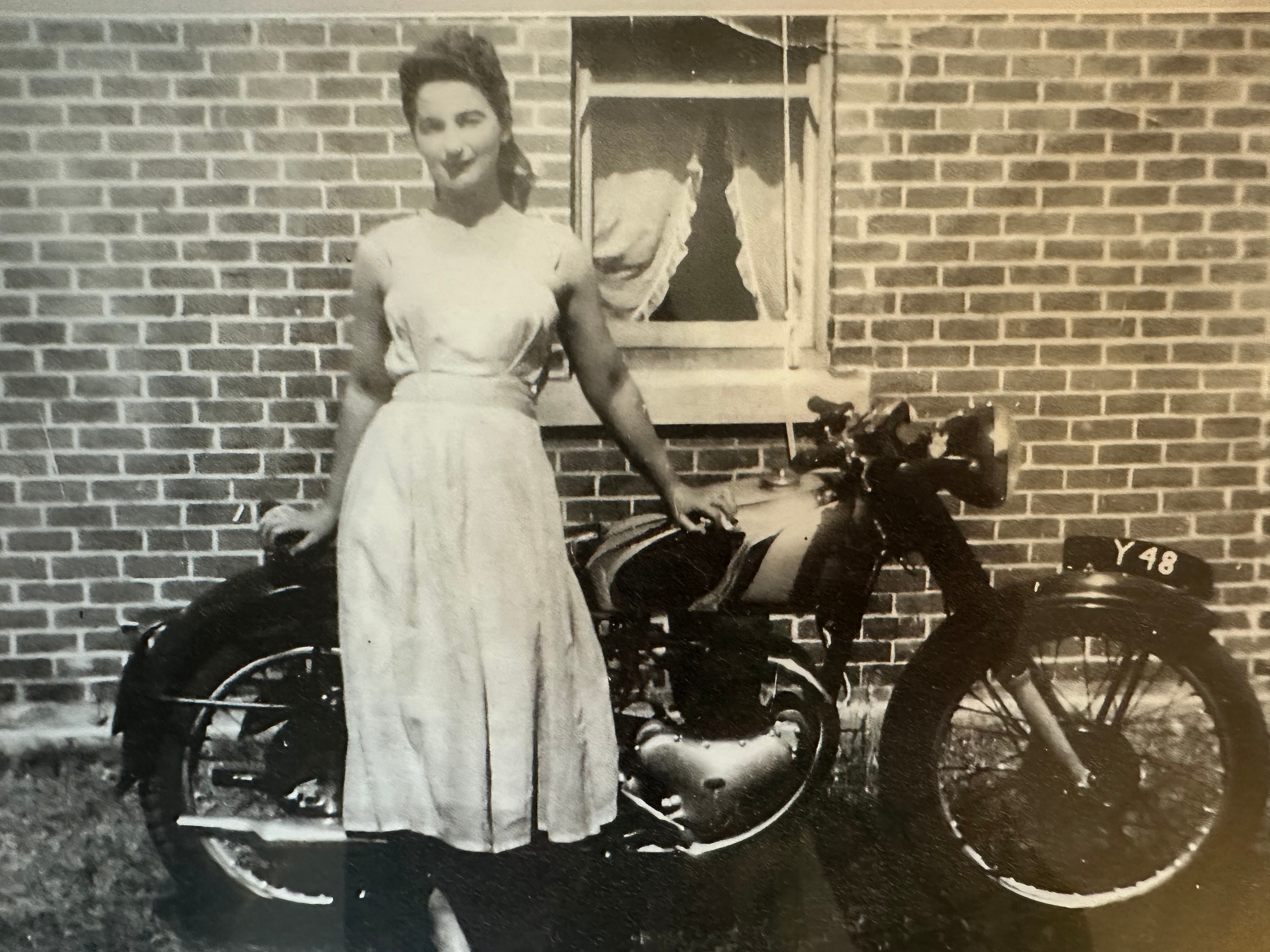 Black and white photo of a young woman in a dress leaning against a motorbike.  