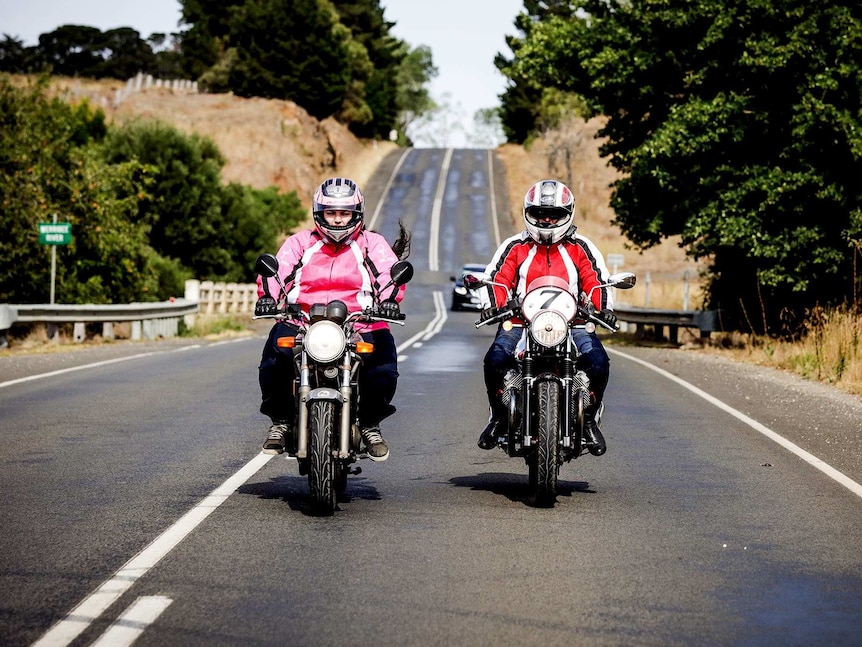 Tracy and Cassie ride their bikes on a country road in leather jackets and helmets