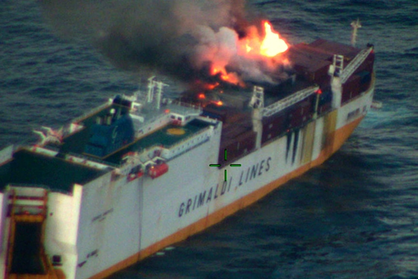 An ship containing oil on fire with flames and billowing grey smoke on the ocean.