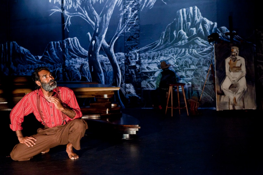 An Indigenous man crouches to the left of stag in front of a dimly lit backdrop