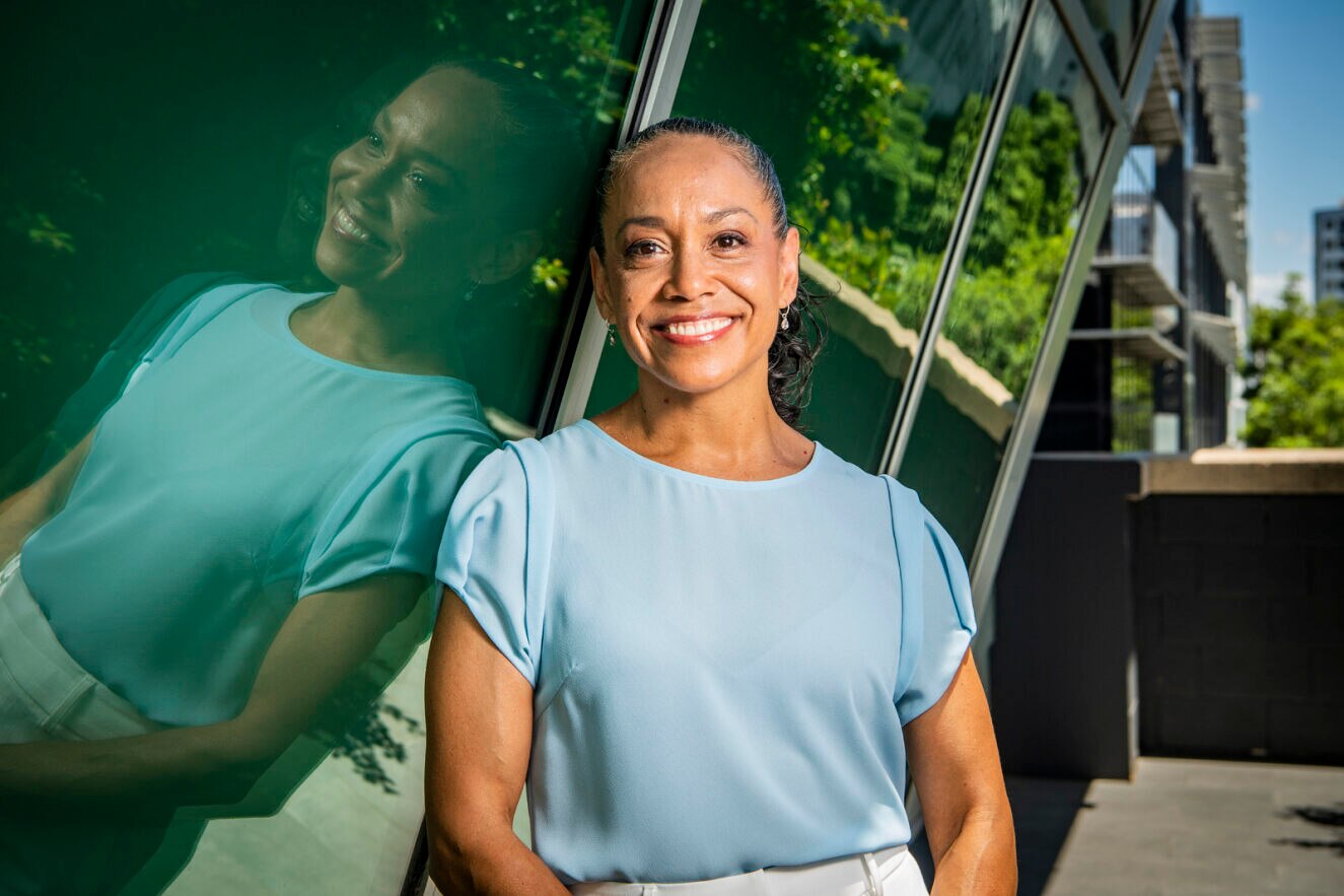 A woman wearing a light blue shirt stands smiling on a balcony.