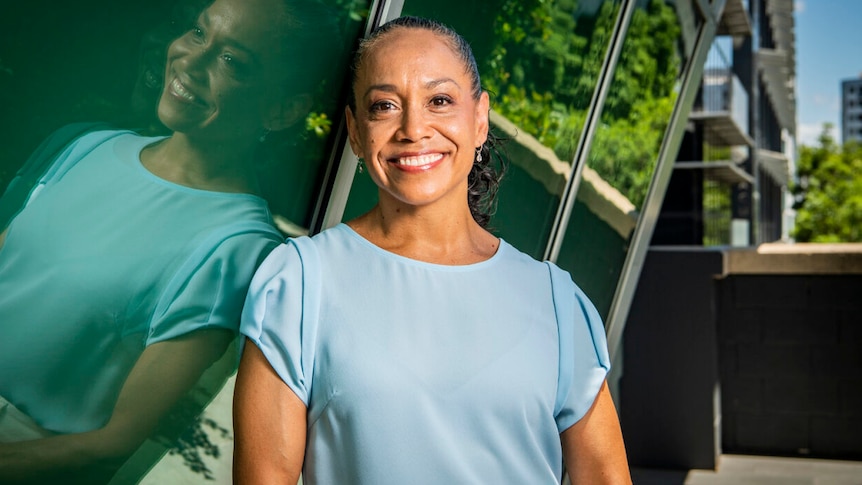 A woman wearing a light blue shirt stands smiling on a balcony.
