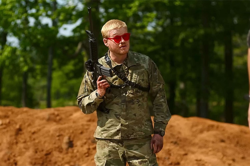 A white man in a camo uniform wears red sunglasses and holds a rifle, standing in front of a mount of dirt and trees.