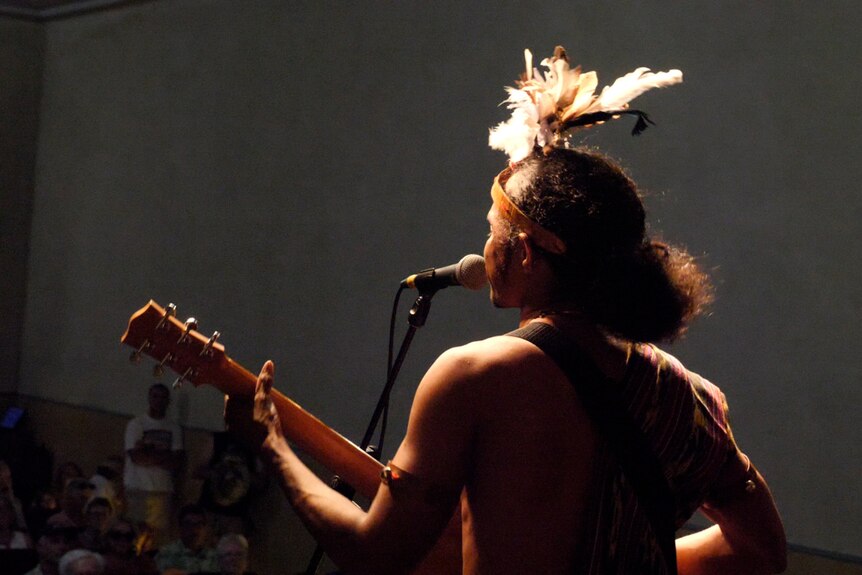 A young man with curly hair, wearing a feather headdress and playing guitar, seen from behind. 