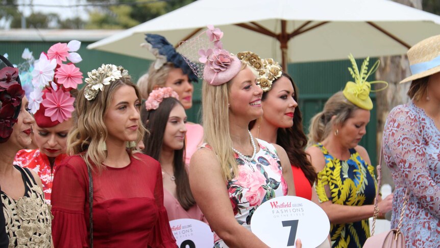 Ladies stand in their racing fashions outdoors, each holding a big number.