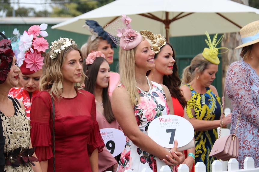 Ladies stand in their racing fashions outdoors, each holding a big number.