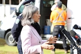 Dr Nicola Spurrier speaking into media microphones at a press conference while a man in the background gets tested for COVID-19