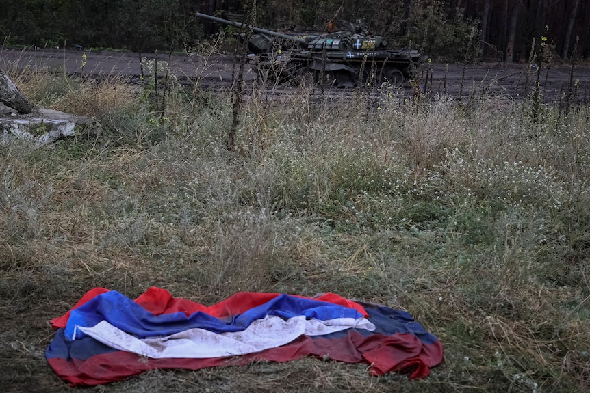 In an area with yellow and green high grass a Russian flag is pictured on the ground. Meters behind it a destroyed Russian tank.