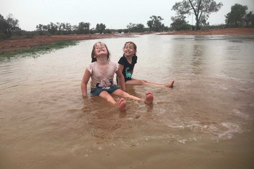 Two young girls sit, smiling, with wet hair, in an expanse of water on open country, as rain falls.